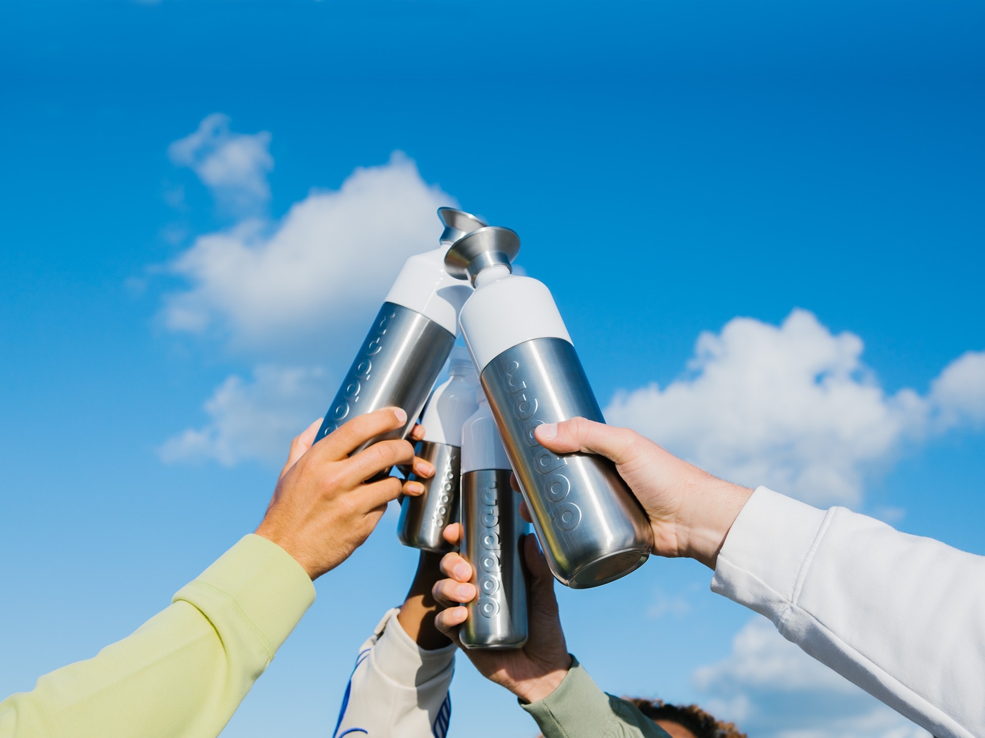 Four different sized Dopper Steel bottles are raised in front of a blue sky